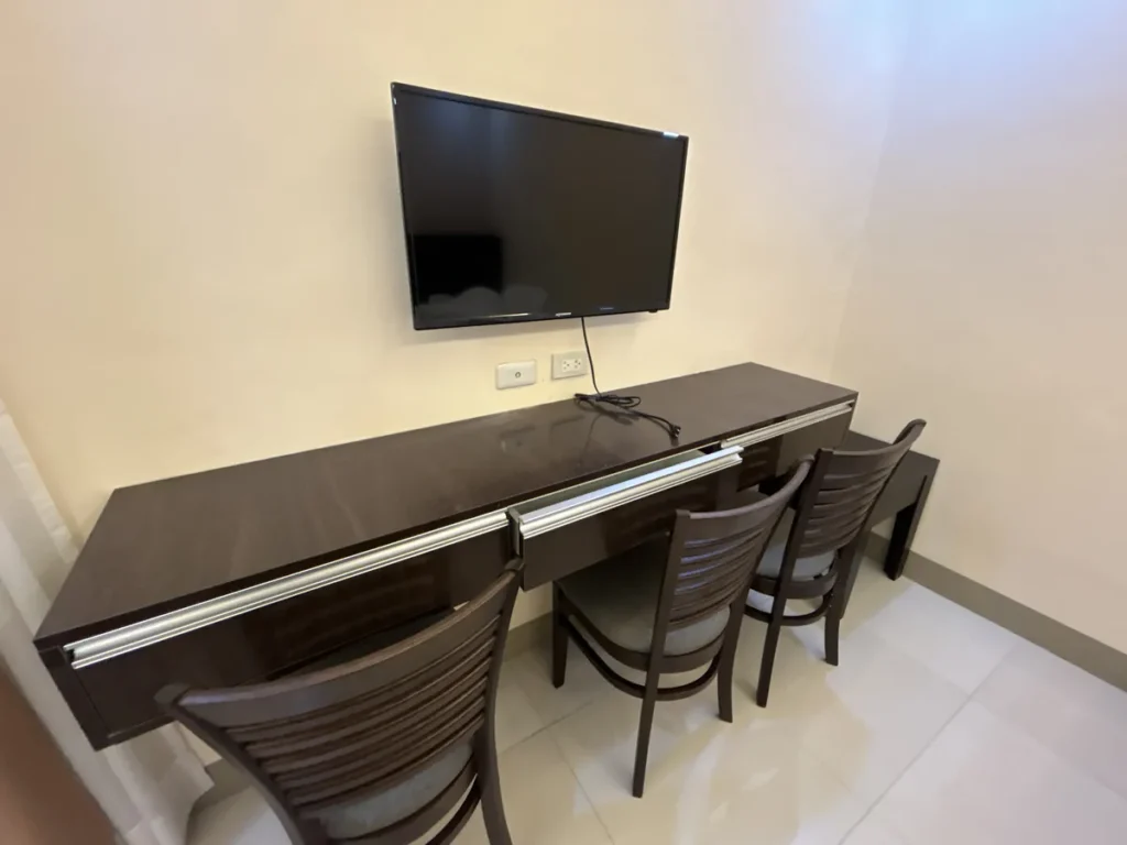 필리핀 세부 CIJ 어학원 외부 기숙사 3인실로, 벽걸이 TV 아래 서랍이 달린 긴 원목 책상과 의자 3개가 나란히 놓인 학습 공간입니다.