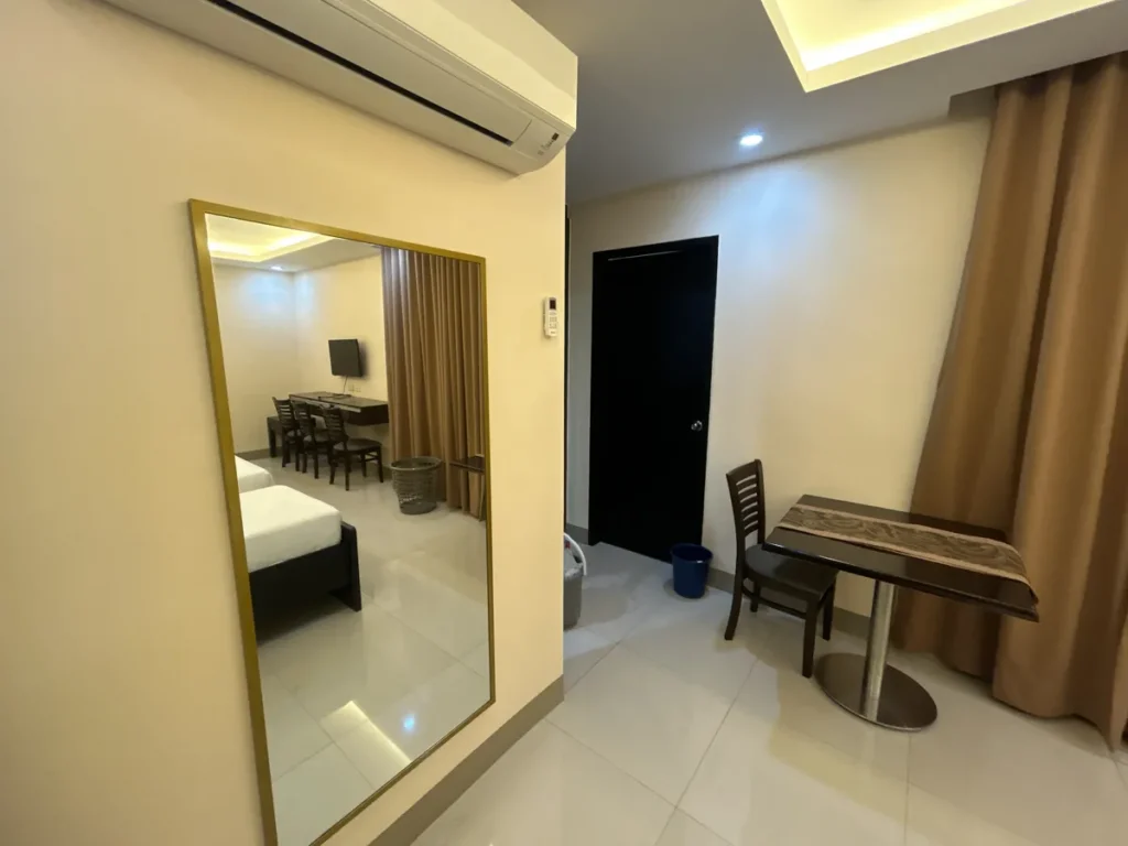 필리핀 세부 CIJ 어학원 외부 기숙사 3인실로, 벽걸이 에어컨 아래 금색 테두리 전신 거울과 창가 쪽 원형 식탁·의자가 놓인 공간입니다.