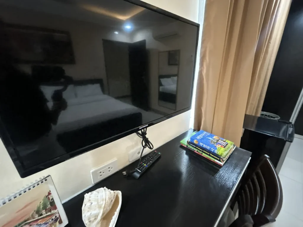 필리핀 세부 CIJ 어학원 외부 기숙사 3인실로, 벽걸이 TV 아래 책상 위에 리모컨과 영어 학습 서적이 놓이고 콘센트가 보이는 모습입니다.