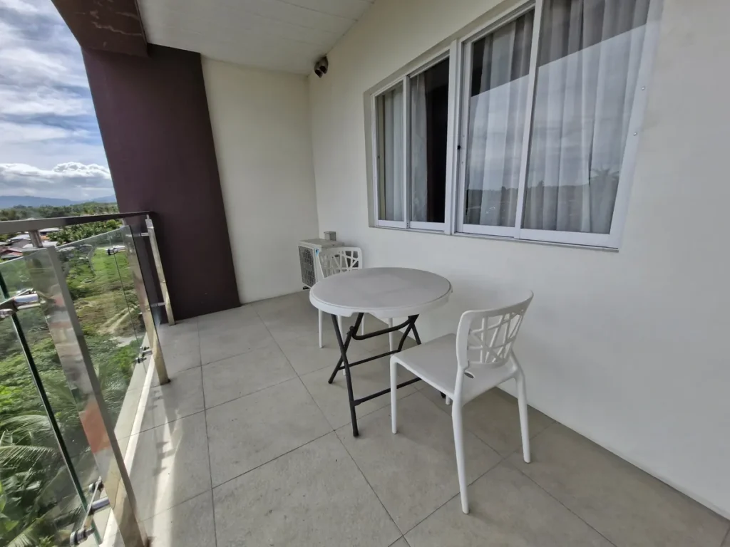 필리핀 세부 CIJ 어학원 외부숙소 공용공간으로, 커튼 달린 창문 옆 발코니에 접이식 원형 테이블과 흰색 의자를 둔 모습입니다.