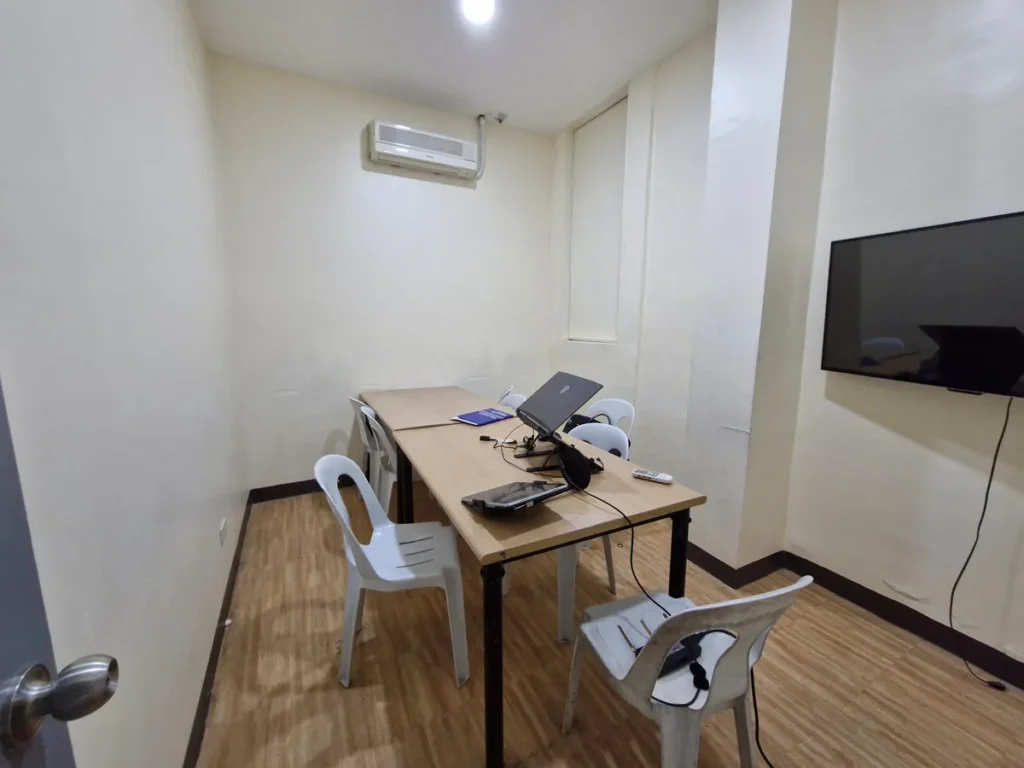 필리핀 세부 CIJ 어학원 그룹 강의실로, 벽걸이 TV와 에어컨이 있고 노트북이 놓인 테이블을 흰색 의자가 둘러싼 교실입니다.