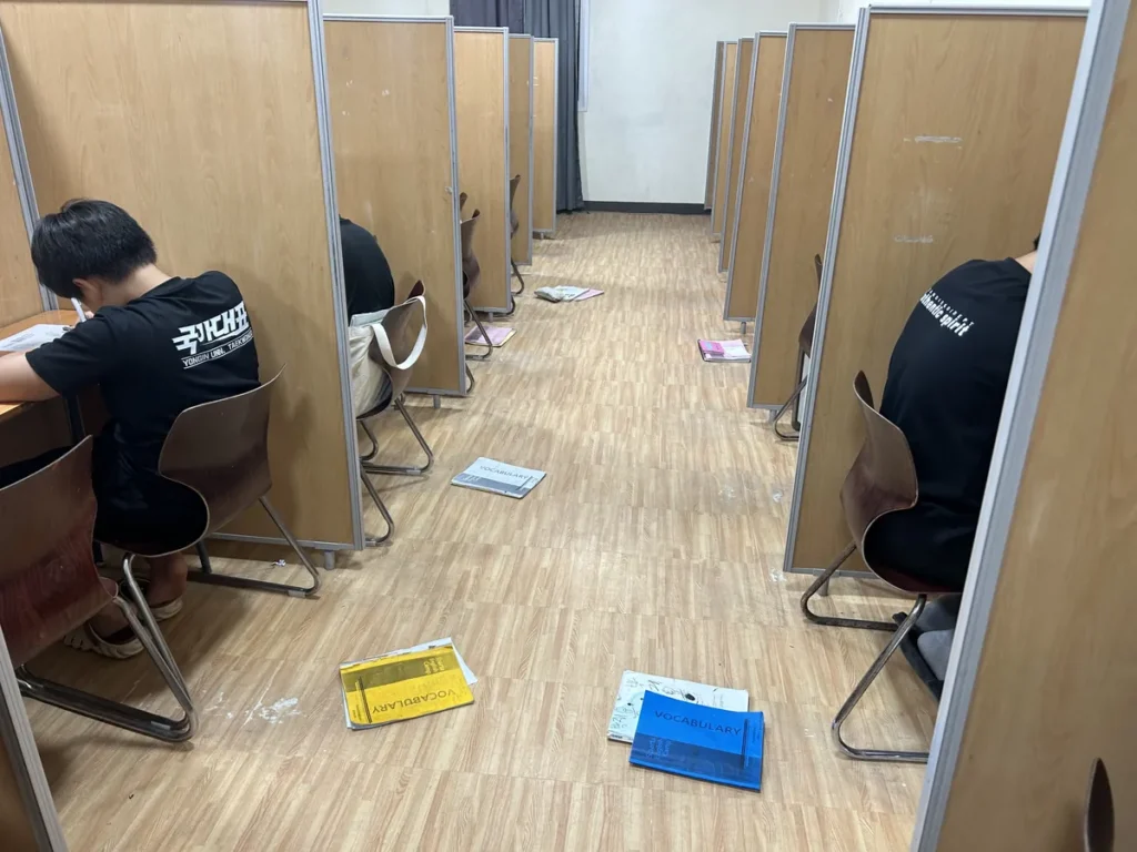 필리핀 세부 CIJ 어학원 자습실로, 양쪽으로 늘어선 칸막이 책상에 학생들이 앉아 공부하고 통로 바닥에 단어 교재가 놓여 있는 모습입니다.
