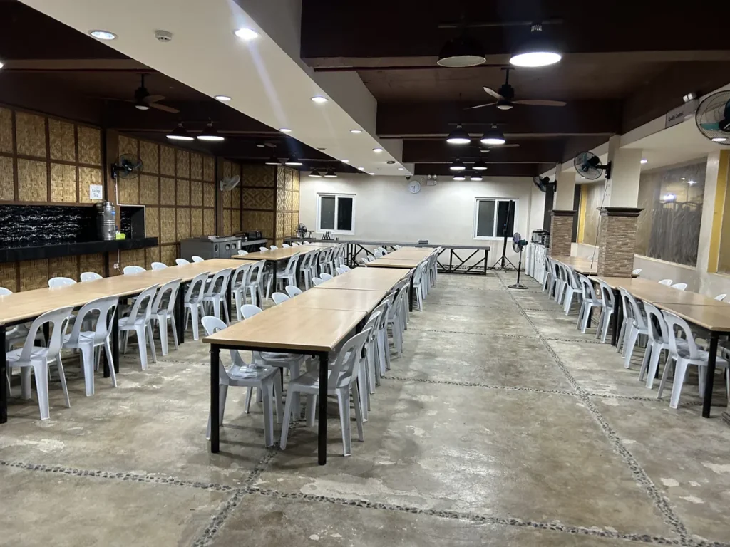 필리핀 세부 CIJ 어학원 식당으로, 저녁 시간 천장 조명과 벽걸이 선풍기가 켜진 가운데 원목 테이블이 정돈된 실내 모습입니다.