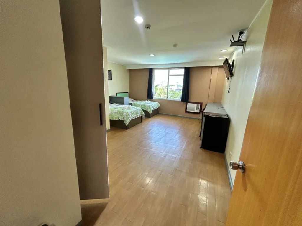 필리핀 세부 시필스(CPILS) 어학원 2인실 프리미엄룸으로, 출입문에서 바라본 긴 마룻바닥 너머 침대 2개와 창문, 오른쪽 냉장고가 보이는 모습입니다.