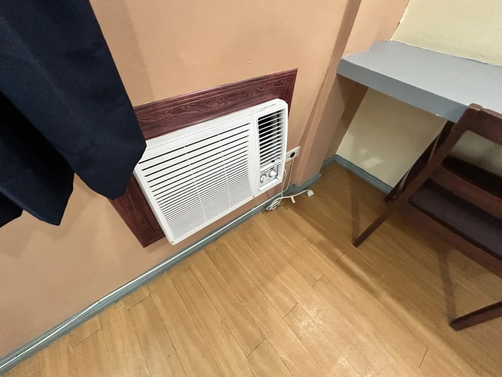 필리핀 세부 시필스(CPILS) 어학원 2인실 프리미엄룸으로, 커튼 옆 벽면에 매립된 창문형 에어컨과 책상·의자 일부가 보이는 모습입니다.