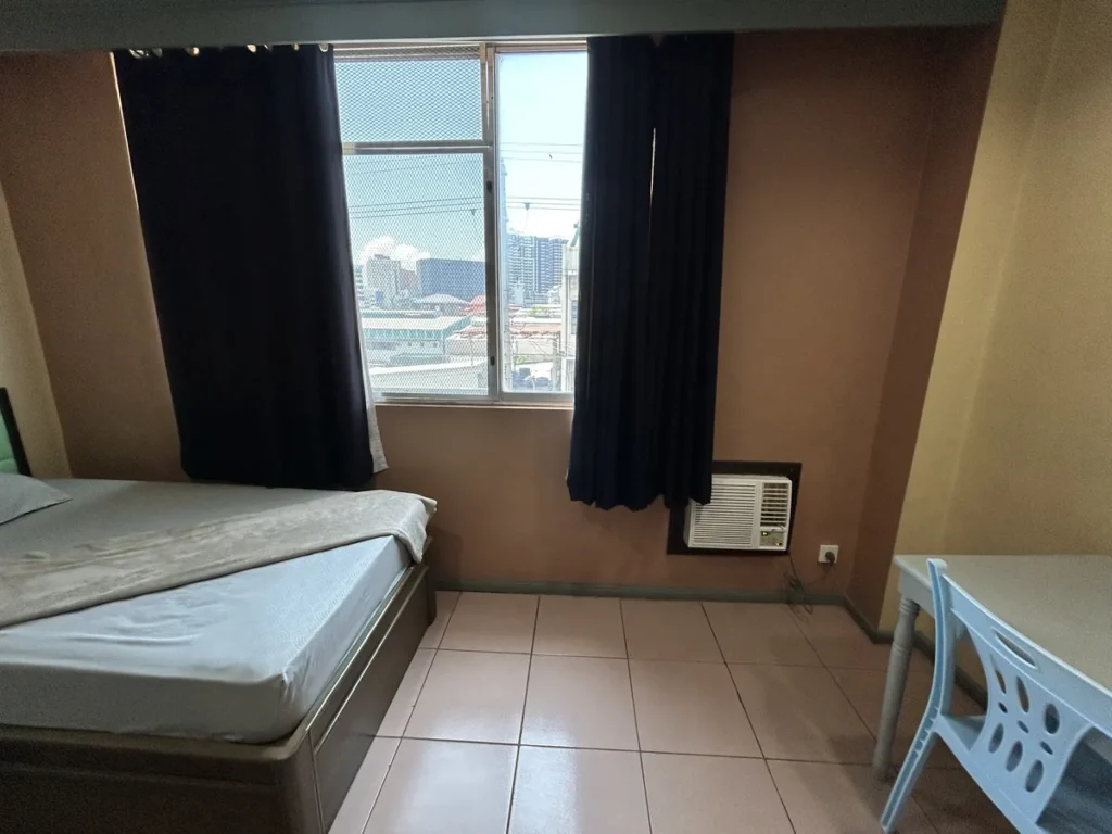 필리핀 세부 시필스(CPILS) 어학원 기숙사 2인실로, 커튼 사이 창밖으로 세부 시내 건물이 보이고 아래에 창문형 에어컨이 설치되어 있습니다.
