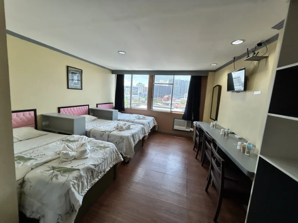 필리핀 세부 시필스(CPILS) 어학원 기숙사 3인실 프리미엄룸으로, 침대 3개와 긴 책상·벽걸이 TV가 놓이고 큰 창으로 시내가 보이는 객실입니다.