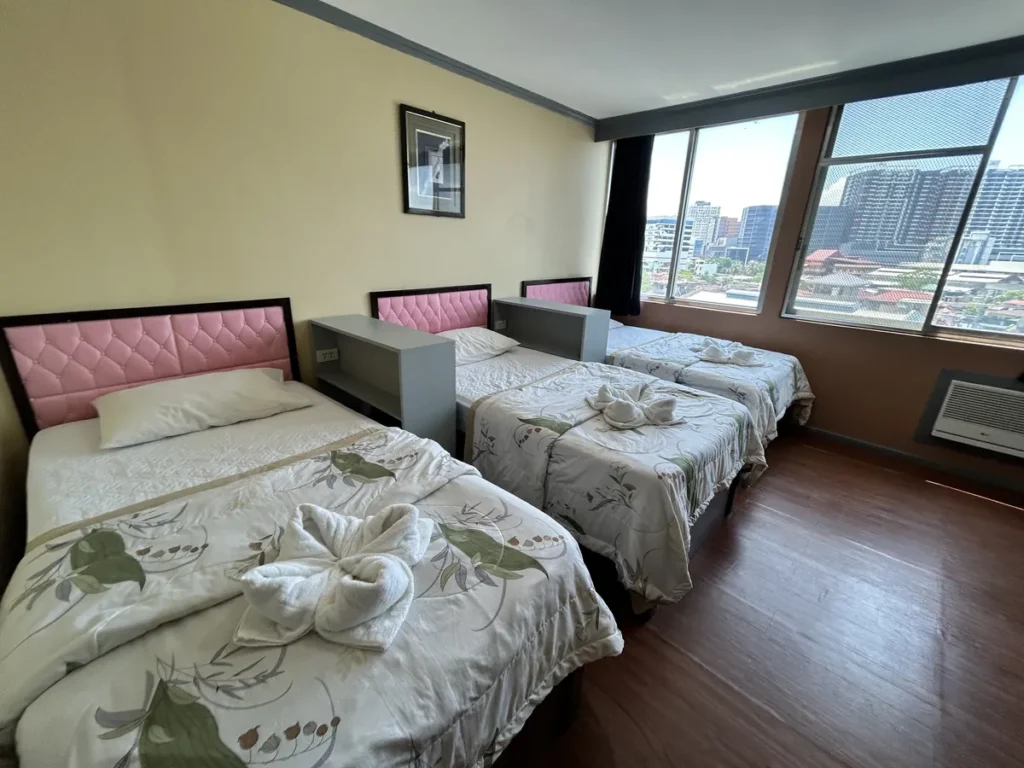 필리핀 세부 시필스(CPILS) 어학원 기숙사 3인실 프리미엄룸으로, 분홍 헤드보드 싱글침대 3개 사이에 협탁이 있고 수건이 올려진 모습입니다.
