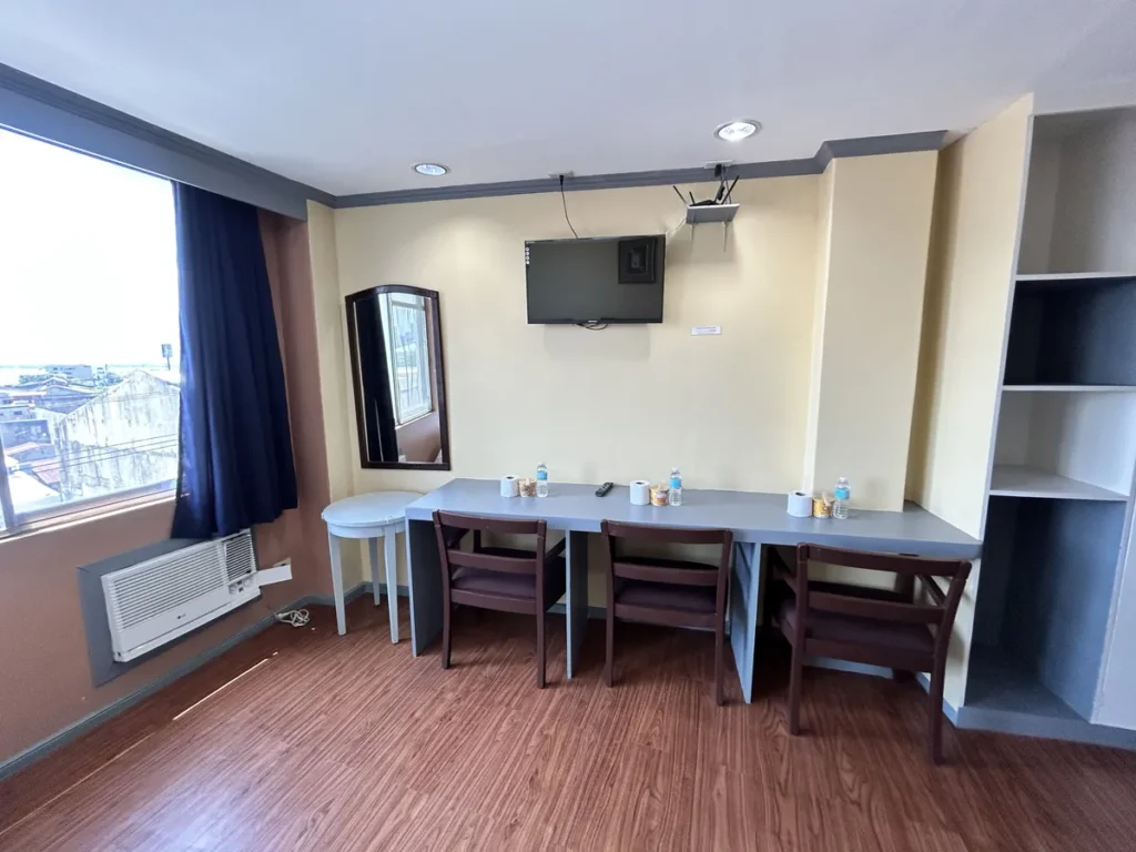 필리핀 세부 시필스(CPILS) 어학원 기숙사 3인실 프리미엄룸으로, 의자 3개가 놓인 긴 책상과 벽걸이 TV·거울·창문형 에어컨이 있는 공간입니다.