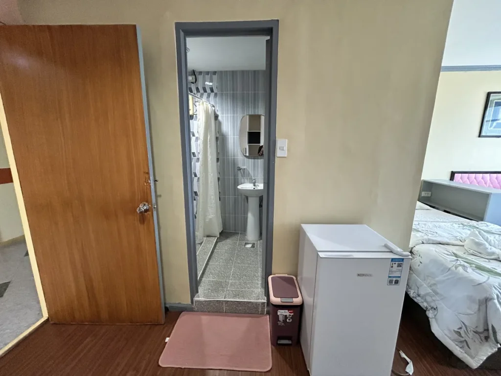 필리핀 세부 시필스(CPILS) 어학원 기숙사 3인실 프리미엄룸으로, 출입문 옆 개별 욕실 입구와 소형 냉장고·휴지통·분홍 발매트가 보이는 모습입니다.
