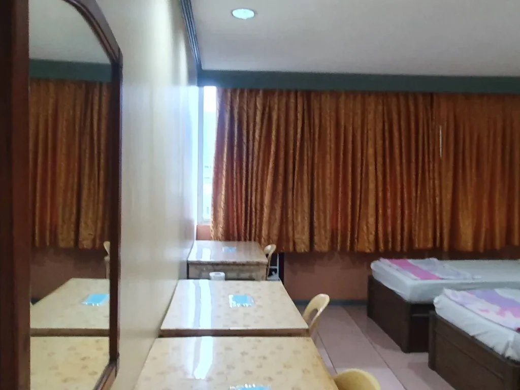 필리핀 세부 시필스(CPILS) 어학원 기숙사 3인실 일반룸으로, 주황빛 커튼이 드리운 창가 쪽에 침대와 책상이 놓인 객실 안쪽 모습입니다.