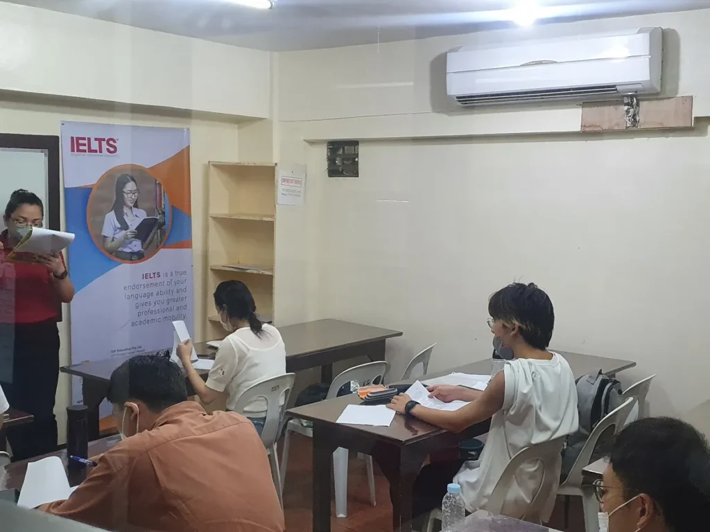 필리핀 세부 시필스(CPILS) 어학원 그룹 강의실로, IELTS 배너가 세워진 교실에서 학생들이 책상에 앉아 자료를 보는 수업 모습입니다.