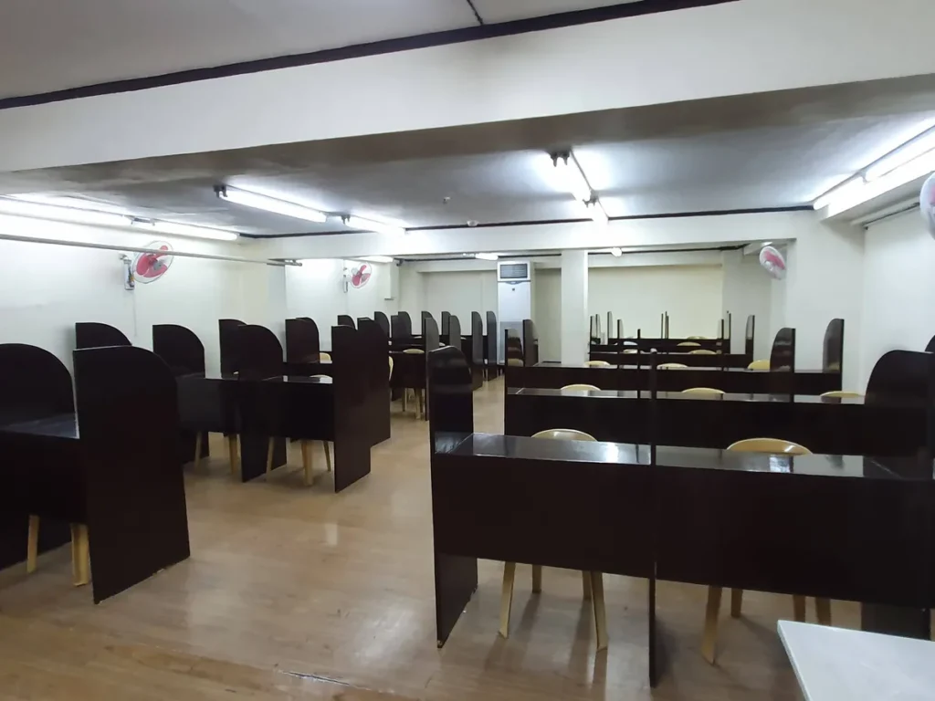 필리핀 세부 시필스(CPILS) 어학원 시험장으로, 칸막이가 있는 짙은 갈색 개인 책상이 여러 줄로 배치된 넓은 실내 모습입니다.