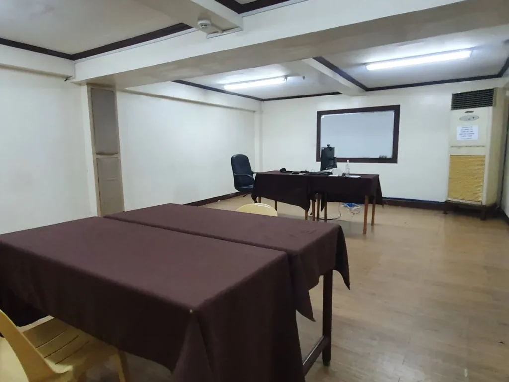 필리핀 세부 시필스(CPILS) 어학원 시험장으로, 천을 덮은 책상들 너머 모니터를 둔 감독관 책상과 화이트보드, 스탠드형 에어컨이 보입니다.