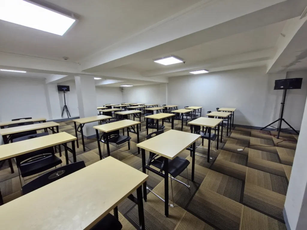 필리핀 세부 시필스(CPILS) 어학원 아이엘츠 시험장으로, 체크무늬 카펫 위 1인용 책상과 의자가 배치되고 양쪽에 스탠드 스피커가 설치된 시험실입니다.