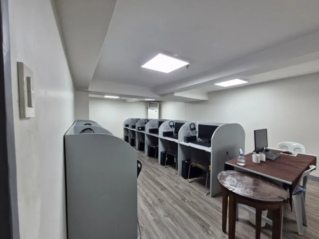필리핀 세부 시필스(CPILS) 어학원 아이엘츠 시험장으로, 컴퓨터 칸막이 부스들과 감독관용 책상·모니터가 함께 놓인 컴퓨터 시험실 전경입니다.