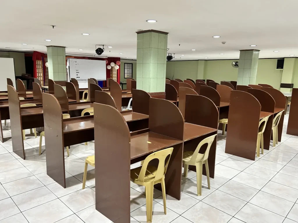 필리핀 세부 시필스(CPILS) 어학원 토익시험장으로, 갈색 칸막이 책상과 플라스틱 의자가 여러 줄로 배치된 시험 공간입니다.