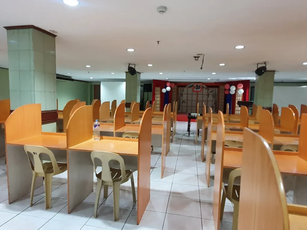필리핀 세부 시필스(CPILS) 어학원 토익시험장으로, 원목 칸막이 책상이 줄지어 있고 안쪽에 풍선으로 꾸민 무대가 보입니다.