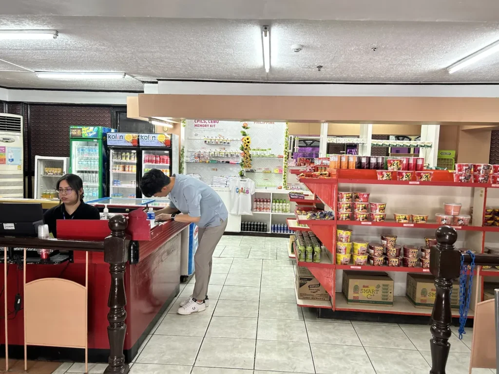 필리핀 세부 시필스(CPILS) 어학원 매점으로, 직원이 있는 계산대 뒤로 음료 냉장고와 기념품 티셔츠·라면 진열대가 놓인 모습입니다.