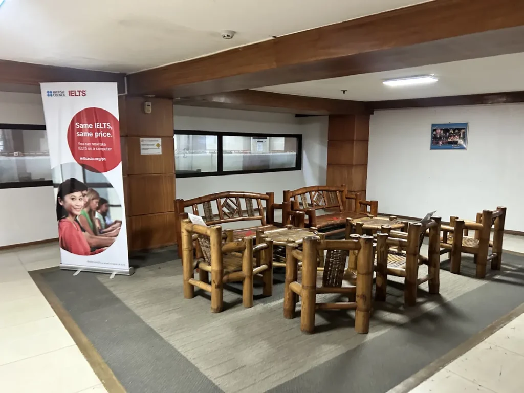 필리핀 세부 시필스(CPILS) 어학원 휴게공간으로, 카펫 위에 대나무 의자와 테이블이 모여 있고 IELTS 안내 배너가 세워져 있습니다.