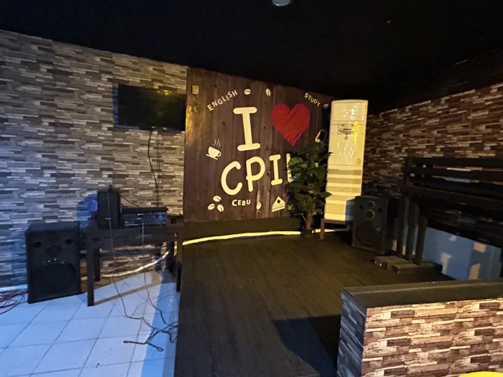 필리핀 세부 시필스(CPILS) 어학원 휴게공간으로, I ♥ CPILS 벽면 장식 앞 낮은 무대와 스피커·TV가 설치된 공연 공간입니다.
