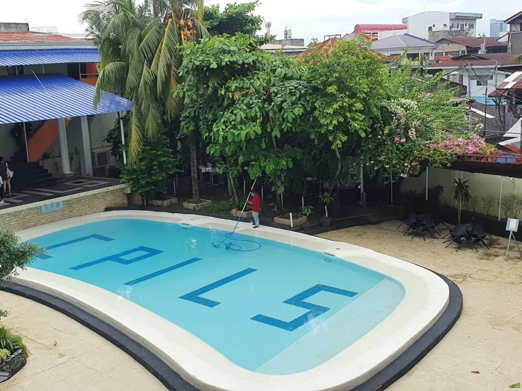 필리핀 세부 시필스(CPILS) 어학원 수영장으로, 야자수와 꽃나무 정원 옆 수영장과 테라스의 검은 의자·테이블이 함께 보입니다.