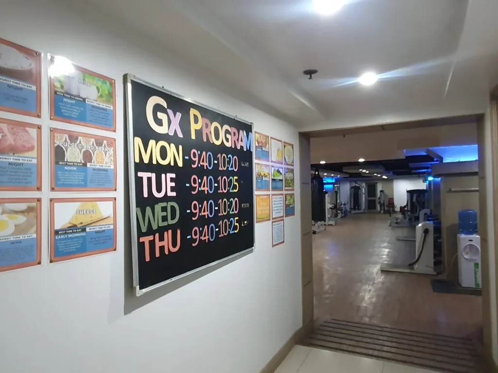 필리핀 세부 시필스(CPILS) 어학원 헬스장 입구로, 요일별 GX 프로그램 시간표와 식단 안내 포스터가 벽에 걸린 복도입니다.
