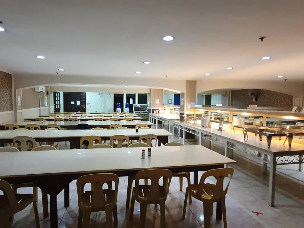 필리핀 세부 시필스(CPILS) 어학원 식당으로, 흰 상판 테이블 줄 옆에 조명이 켜진 배식대와 정수기가 보이는 실내입니다.
