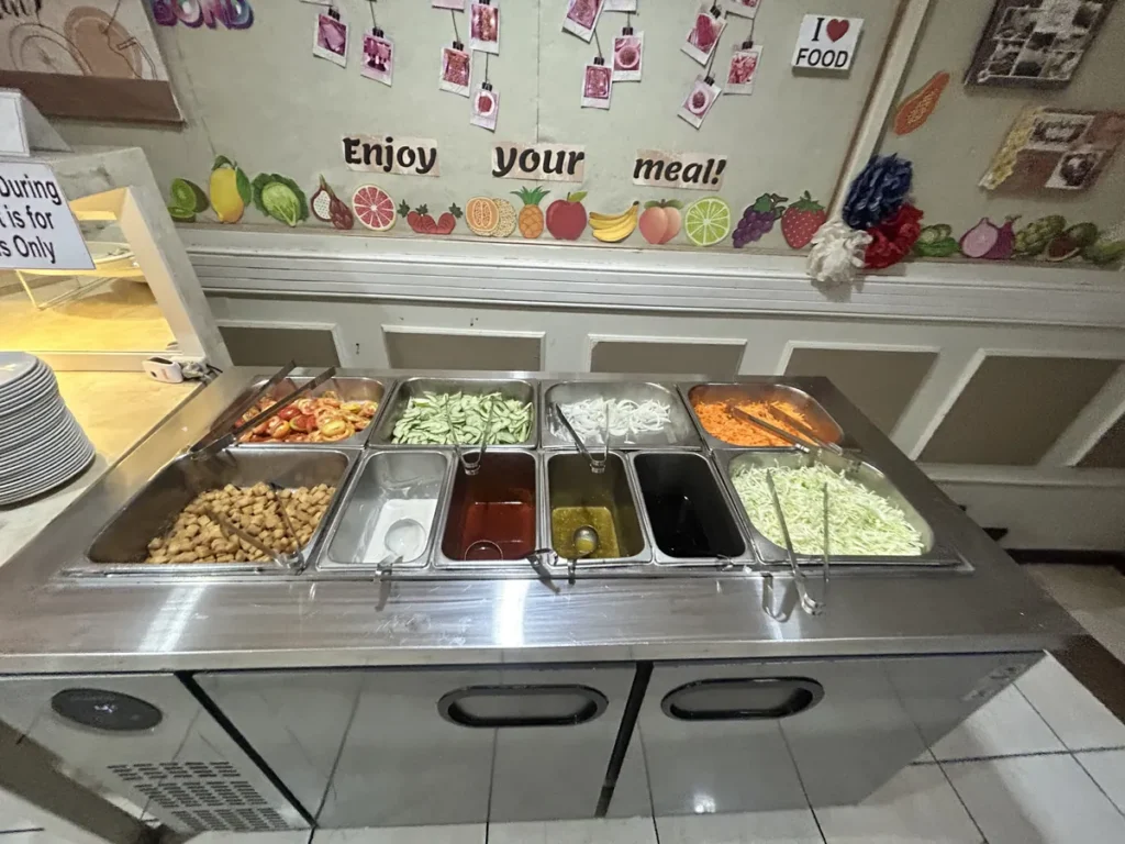 필리핀 세부 시필스(CPILS) 어학원 식당 식사 메뉴로, 과일 벽장식 아래 토마토·오이·양배추와 드레싱이 칸칸이 담긴 샐러드바입니다.