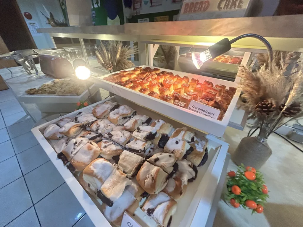 필리핀 세부 시필스(CPILS) 어학원 식당 식사 메뉴로, 조명 아래 캐러멜 브레드푸딩과 크림 올린 빵이 2단 진열대에 놓인 디저트 코너입니다.